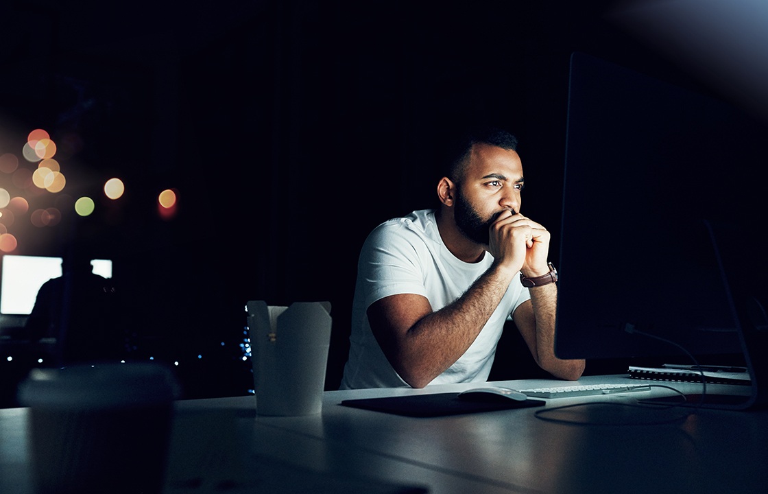 Homme dans le noir, les coudes sur son bureau et appuyant la tête sur ses mains, illuminé par son écran d'ordinateur non visible