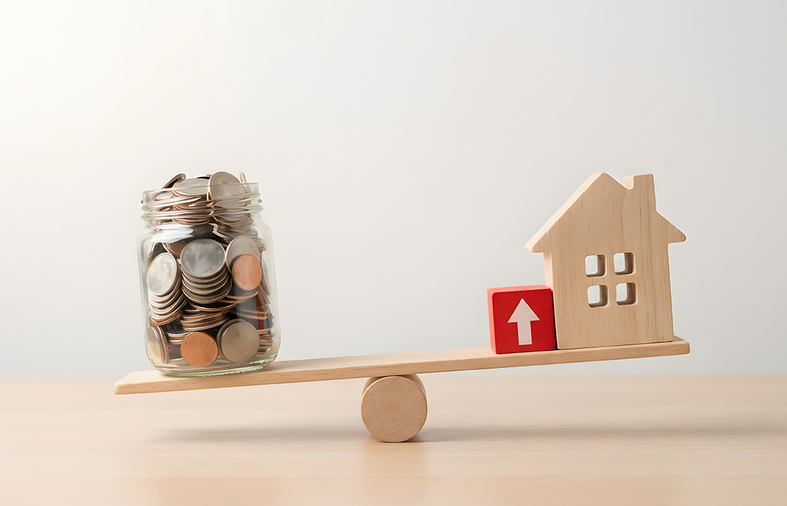 balance board with a jar of coins on one side and a wooden house on the other