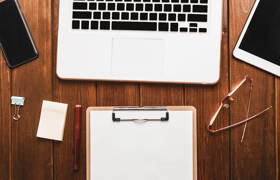 wooden table with white laptop, tablet, mobile phone, glasses, clipboard, sticky notes, pen and paper clip on it