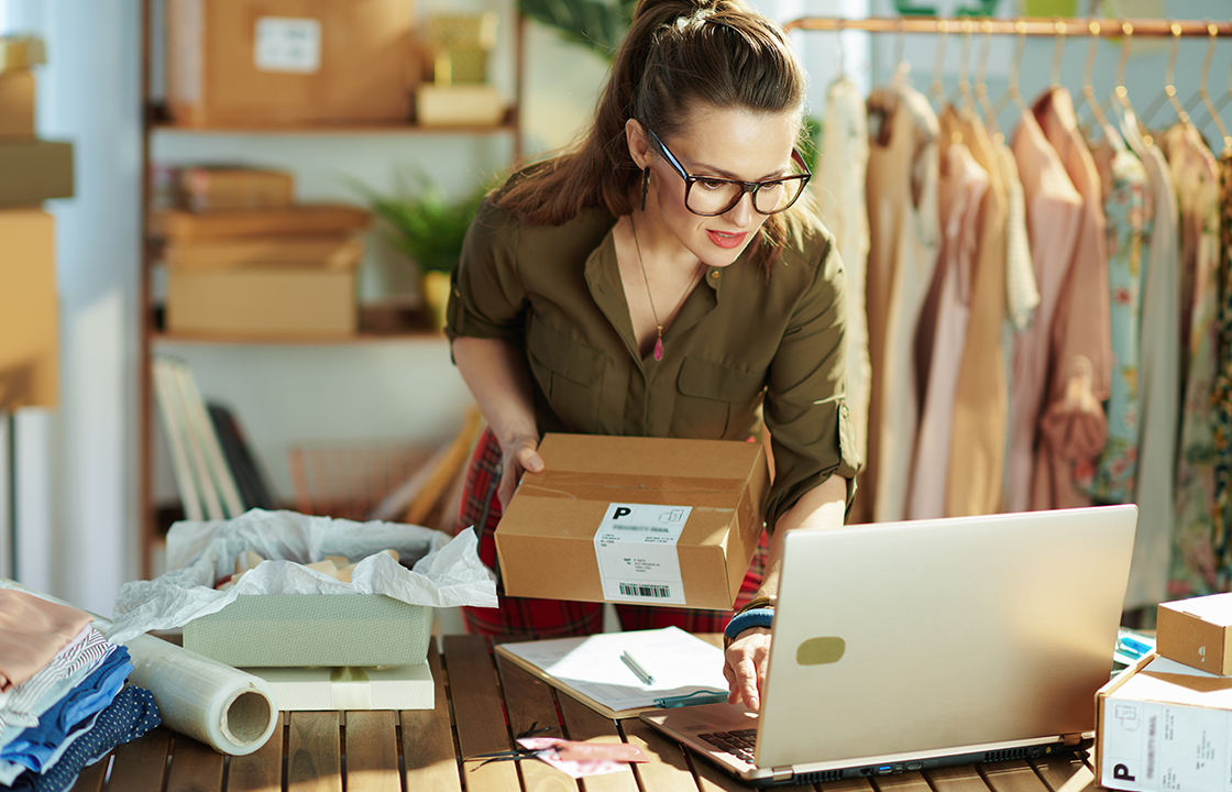 woman at a laptop with a rack of clothes behind her and shipping boxes around